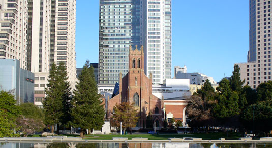 Red stone church near green trees, surrounded by skyscrapers.